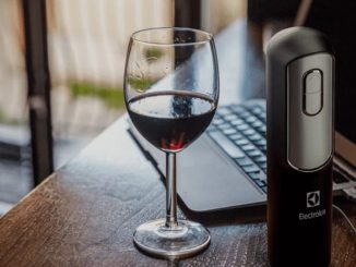 Abridor de vinhos elétrico Electrolux sendo recarregado via USB em um notebook, com uma taça de vinho ao lado, sobre uma mesa.