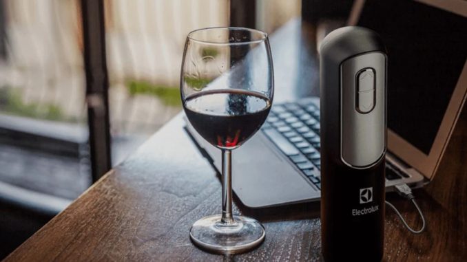 Abridor de vinhos elétrico Electrolux sendo recarregado via USB em um notebook, com uma taça de vinho ao lado, sobre uma mesa.