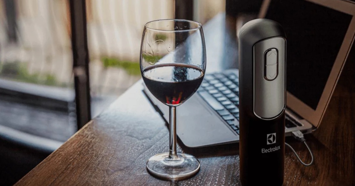 Abridor de vinhos elétrico Electrolux sendo recarregado via USB em um notebook, com uma taça de vinho ao lado, sobre uma mesa.